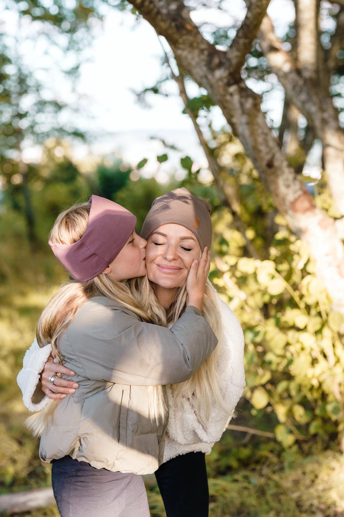 En dotter som pussar sin mamma på kinden. Dottern bär ett pannband från OICAN och mamman bär en SafeHat Taupe. Det är soligt och de befinner sig i skogen.