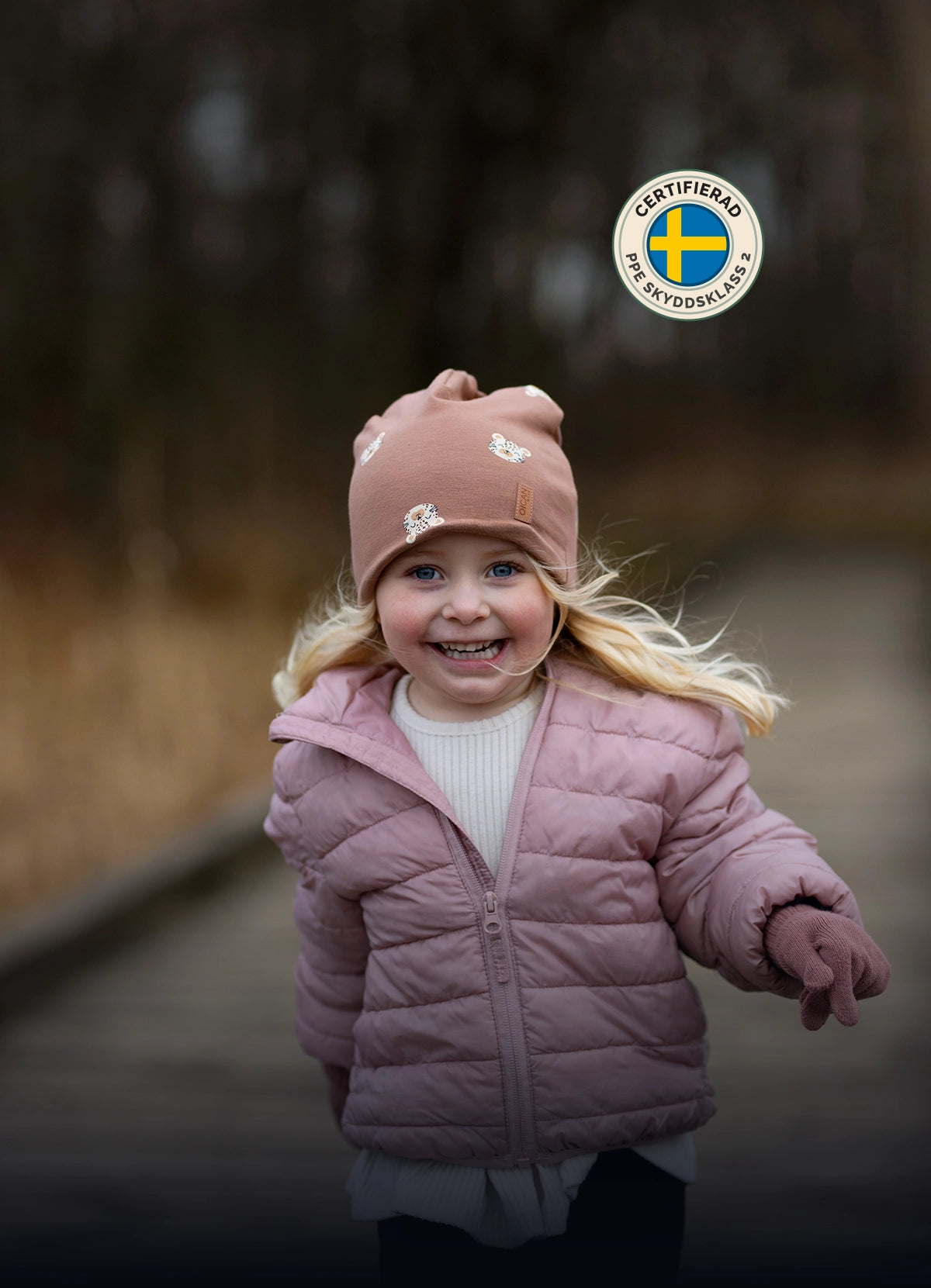 Ung tjej i rosa dunjacka och fingervantar springer på en brygga. Flickan har på sig OICAN safehat i ljusbrunt med små leopader. Bilder har även en Svensk certifierad logga för PPE Skyddsklass 2